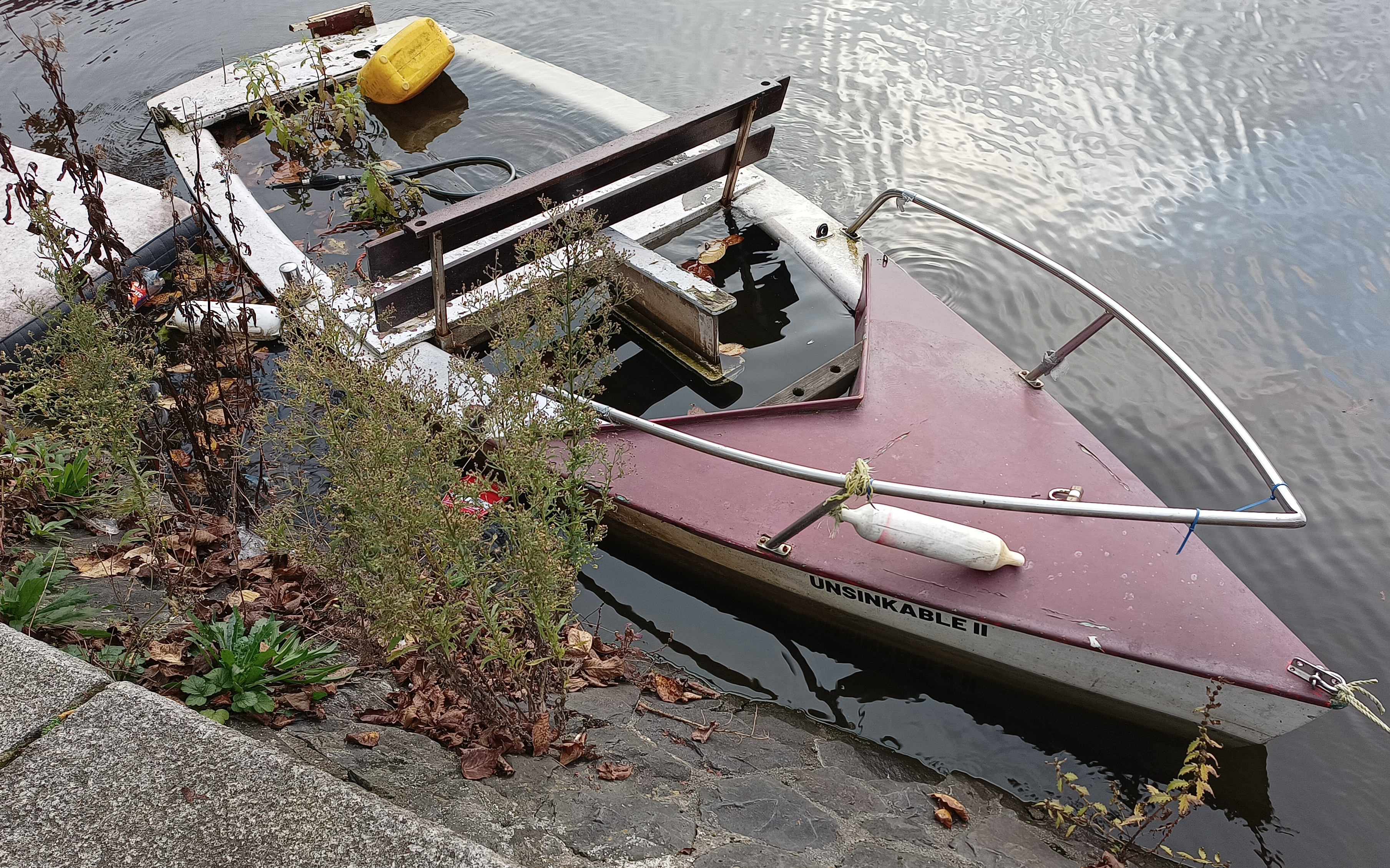 A boat docked in an Amsterdam canal. There are plants growing inside, and it's filled to the brim with water. The name of the boat is Unsinkable II. Taken November 2024.